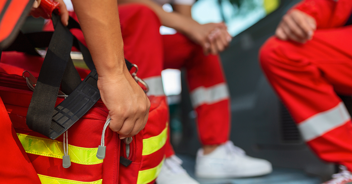 Auf dem Bild sieht man drei Personen in roten Einsatzanzügen mit reflektierenden Streifen – typische Kleidung von Rettungskräften oder Sanitätern. Zwei sitzen, eine Person hält eine große, rote Notfalltasche mit gelben Reflexstreifen und schwarzen Gurten in der Hand. Die Szene wirkt ruhig, vermutlich in einer kurzen Pause oder vor dem nächsten Einsatz. Im Hintergrund ist ein unscharfer Innenraum eines Fahrzeugs zu erkennen, wahrscheinlich ein Rettungswagen. Die Stimmung ist konzentriert und bereit.