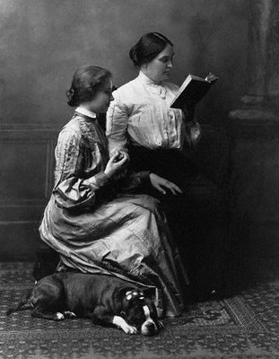 Das Schwarz-Weiß-Foto zeigt zwei Frauen und einen Hund in einem Innenraum mit Teppichboden. Die Szene wirkt ruhig und intim und stammt vermutlich aus dem späten 19. oder frühen 20. Jahrhundert.
Links sitzt eine Frau mit hellem Gesicht und hochgestecktem Haar auf dem Boden oder einem niedrigen Hocker. Sie trägt ein bodenlanges, elegantes Kleid mit langen Ärmeln. Ihre Haltung ist aufrecht, und sie richtet ihre Aufmerksamkeit nach rechts zur zweiten Frau, die etwas höher sitzt – vermutlich auf einem Stuhl.
Die zweite Frau, rechts im Bild, trägt ebenfalls ein langes Kleid oder Rock mit einer weißen Bluse. Sie hat dunkle, hochgesteckte Haare und liest konzentriert aus einem Buch. Das Buch hält sie mit beiden Händen etwas vor ihr Gesicht.
Zu Füßen der beiden Frauen liegt ein großer Hund – wahrscheinlich ein Boxer – mit dunklem Fell und heller Schnauze. Er liegt entspannt auf dem Teppich und scheint zu schlafen.
Die gesamte Szene strahlt Ruhe, Bildung und eine enge Verbindung zwischen den beiden Frauen aus – möglicherweise eine Lehrsituation oder ein Moment gemeinsamen Lesens.
