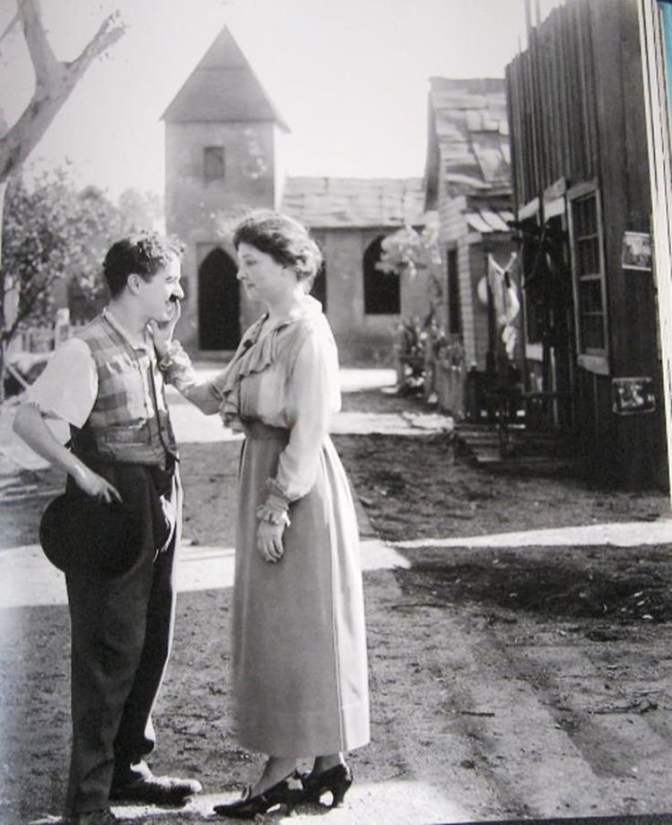 Das Schwarz-Weiß-Foto zeigt zwei Menschen, die sich einander zugewandt gegenüberstehen – eine Frau und ein Mann. Die Szene spielt im Freien auf einer staubigen Straße, die durch ein kleines Dorf oder eine Western-Kulisse führt. Im Hintergrund stehen einfache Gebäude aus Holz sowie ein Haus mit Turm und spitzem Dach. Die Umgebung wirkt ruhig, fast wie eine Filmszene. Die Frau steht rechts im Bild. Sie ist mittleren Alters, trägt ein langes, helles Kleid mit langen Ärmeln und eine Schleife am Ausschnitt. Ihre dunklen Haare sind hochgesteckt. Sie hält mit ihrer rechten Hand das Gesicht des Mannes zärtlich an der Wange und scheint ihn zu „lesen“ – vermutlich mit Hilfe der Berührung, wie es bei taubblinden Menschen üblich ist. Ihr Ausdruck ist sanft und konzentriert. Der Mann links im Bild trägt eine dunkle Hose, ein helles Hemd mit aufgekrempelten Ärmeln und eine gemusterte Weste. Er hält einen schwarzen Hut in der linken Hand und lächelt leicht. Sein Gesichtsausdruck wirkt offen und freundlich, und er steht still, während die Frau ihn berührt. Die Szene strahlt Respekt, Neugier und ein stilles, intensives Miteinander aus.