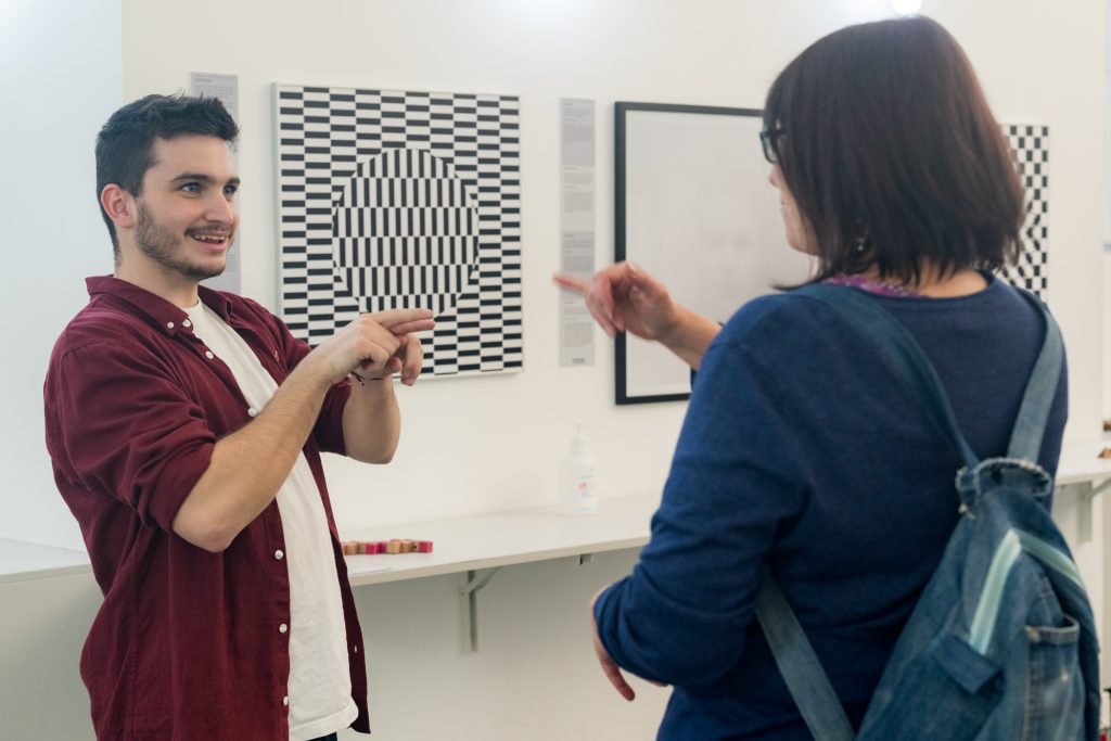 Zwei Menschen unterhalten sich in einem hellen Raum, vermutlich einer Galerie oder einem Museum. Der junge Mann links im Bild hat kurze dunkle Haare, trägt ein weißes T-Shirt und darüber ein offenes bordeauxrotes Hemd. Er lächelt freundlich und kommuniziert mit der Frau gegenüber durch Gebärdensprache. Beide zeigen mit den Fingern, ihre Hände sind erhoben und aufeinander gerichtet.
Die Frau steht rechts im Bild, ihr Gesicht ist nur teilweise sichtbar. Sie trägt eine Brille, ein blaues Oberteil und einen Rucksack aus Jeansstoff. Ihre Haare sind schulterlang und braun.
Im Hintergrund hängen an der weißen Wand moderne Kunstwerke – eines davon ist ein optisches Täuschungsbild in Schwarz-Weiß mit einem Muster aus vertikalen und horizontalen Linien, in dessen Mitte ein Kreis erkennbar ist. Darunter steht eine Flasche mit Desinfektionsmittel auf einem weißen Regal.
Die Szene wirkt freundlich und kommunikativ – beide Personen sind in einem intensiven, aber positiven Austausch miteinander.