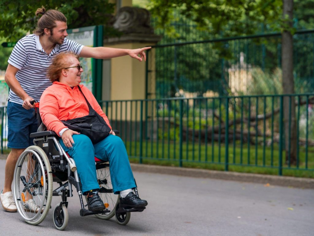 Ein persönlicher Assistent schiebt eine Frau im Rollstuhl. Die beiden haben Spaß.